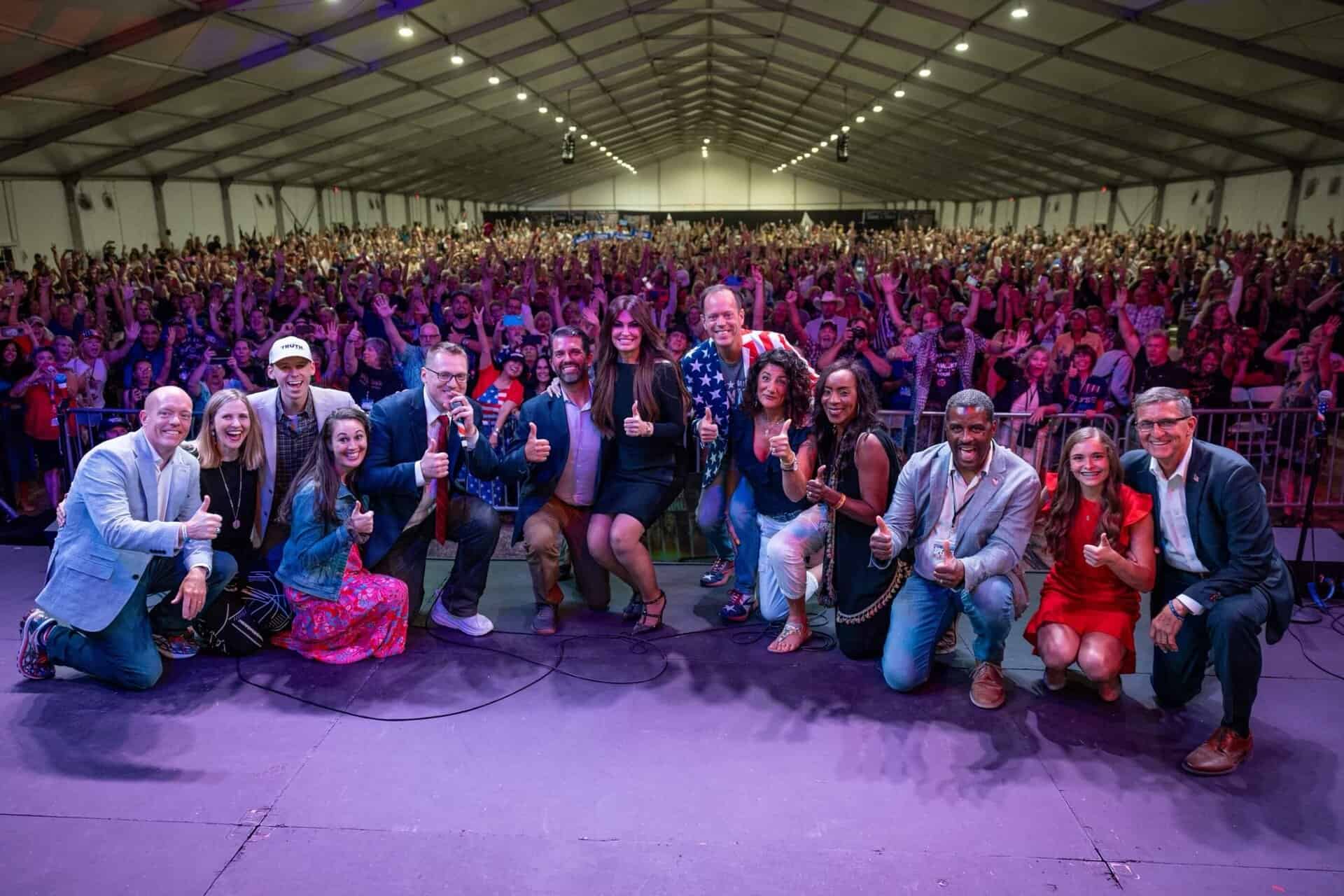 Large group of diverse people on stage with enthusiastic audience in exhibition hall.