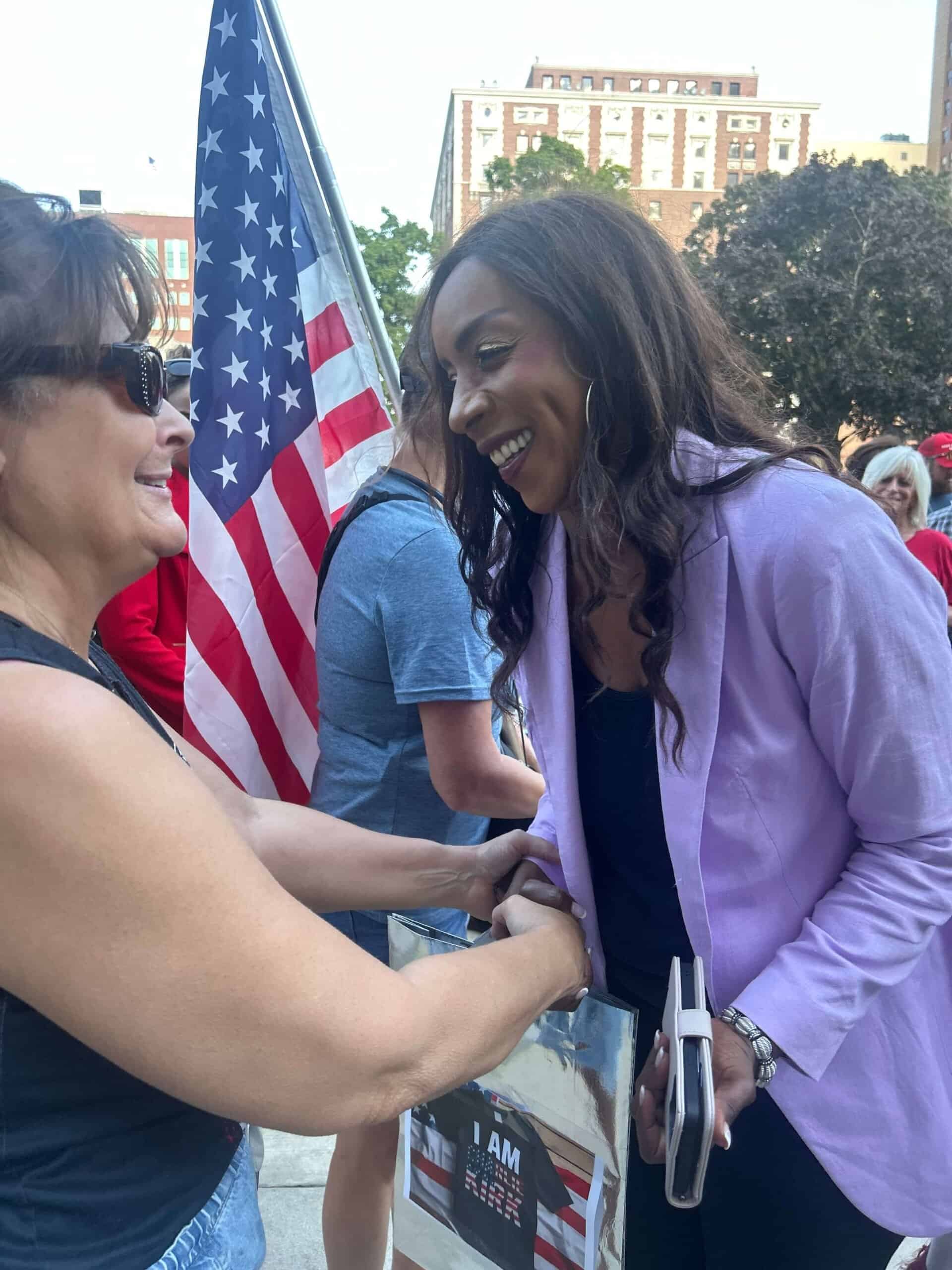 Supportive woman shaking hands with smiling woman at patriotic event, American flag in background.